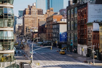 A city street scene features a mixture of modern and older brick buildings. There are large signs, including one advertising retail space for lease. The street is wide and relatively empty, with a few vehicles visible, including a yellow taxi. Architecture includes fire escapes and various styles of windows, with some greenery along the sidewalks.