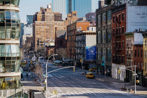 A city street scene features a mixture of modern and older brick buildings. There are large signs, including one advertising retail space for lease. The street is wide and relatively empty, with a few vehicles visible, including a yellow taxi. Architecture includes fire escapes and various styles of windows, with some greenery along the sidewalks.