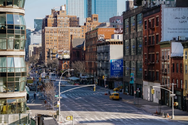 A city street scene features a mixture of modern and older brick buildings. There are large signs, including one advertising retail space for lease. The street is wide and relatively empty, with a few vehicles visible, including a yellow taxi. Architecture includes fire escapes and various styles of windows, with some greenery along the sidewalks.