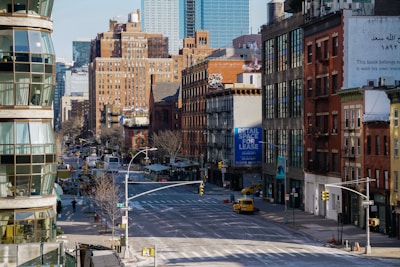 A city street scene features a mixture of modern and older brick buildings. There are large signs, including one advertising retail space for lease. The street is wide and relatively empty, with a few vehicles visible, including a yellow taxi. Architecture includes fire escapes and various styles of windows, with some greenery along the sidewalks.