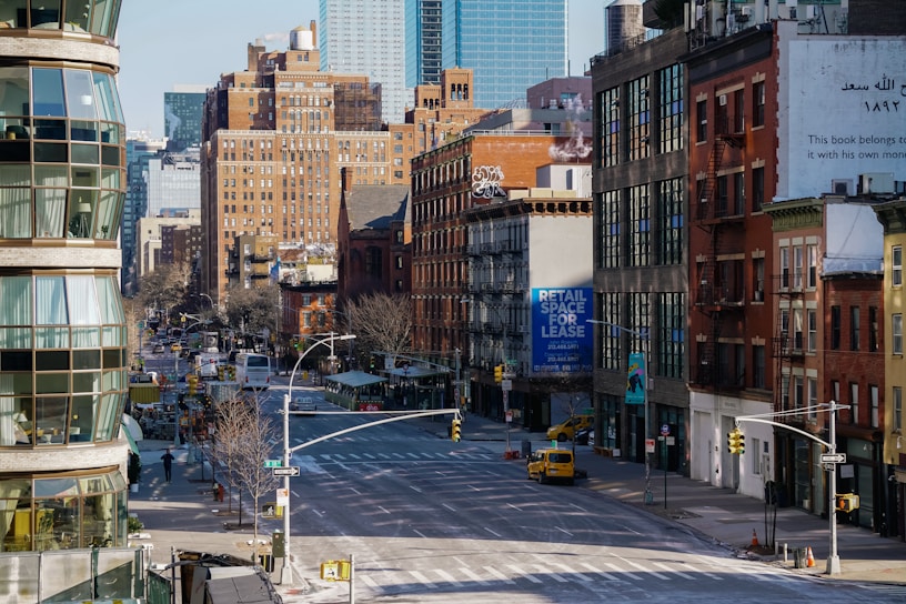 A city street scene features a mixture of modern and older brick buildings. There are large signs, including one advertising retail space for lease. The street is wide and relatively empty, with a few vehicles visible, including a yellow taxi. Architecture includes fire escapes and various styles of windows, with some greenery along the sidewalks.