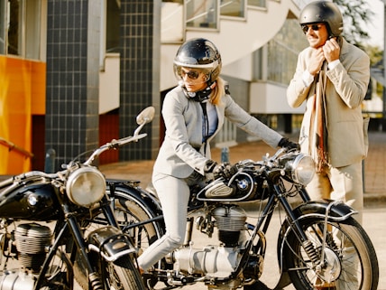 A person in a suit and helmet is seated on a vintage BMW motorcycle, accompanied by another person standing beside them, also in a suit and wearing a helmet. They appear to be outdoors near a modern building with large windows.