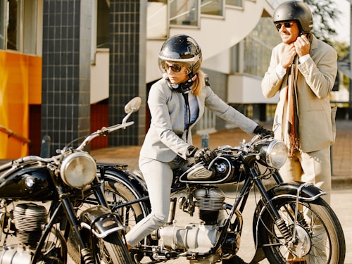 A person in a suit and helmet is seated on a vintage BMW motorcycle, accompanied by another person standing beside them, also in a suit and wearing a helmet. They appear to be outdoors near a modern building with large windows.