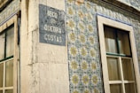 A corner of a building featuring traditional Portuguese ceramic tiles with intricate geometric and floral patterns in blue, yellow, and green. The building has two windows with stone frames and a plaque on the wall displaying the name 'Beco do Quebra Costas'. The tiles and architectural details suggest an old, possibly historical, urban setting.