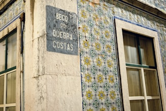 A corner of a building featuring traditional Portuguese ceramic tiles with intricate geometric and floral patterns in blue, yellow, and green. The building has two windows with stone frames and a plaque on the wall displaying the name 'Beco do Quebra Costas'. The tiles and architectural details suggest an old, possibly historical, urban setting.