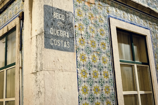 A corner of a building featuring traditional Portuguese ceramic tiles with intricate geometric and floral patterns in blue, yellow, and green. The building has two windows with stone frames and a plaque on the wall displaying the name 'Beco do Quebra Costas'. The tiles and architectural details suggest an old, possibly historical, urban setting.