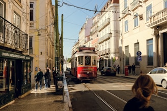 A vintage red tram travels along a narrow street lined with historic European buildings. The street is bustling with people walking and standing around, some near a bookstore with a green facade. The atmosphere suggests an urban setting in Europe.