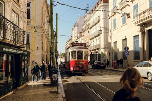A vintage red tram travels along a narrow street lined with historic European buildings. The street is bustling with people walking and standing around, some near a bookstore with a green facade. The atmosphere suggests an urban setting in Europe.