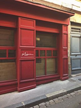 A red storefront with large window panels and the words 'je t'aime' written on one of the doors in a cursive style. The setting appears to be a charming street with cobblestones and a weathered stone facade next to the red structure. The doors and windows have a traditional design, creating a quaint and romantic atmosphere.