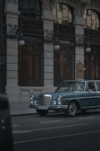 A vintage blue Mercedes-Benz is parked on a city street in front of a luxury boutique store with ornate ironwork and tall windows. The lighting is subdued, conveying a sense of elegance and timelessness.