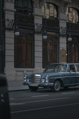 A vintage blue Mercedes-Benz is parked on a city street in front of a luxury boutique store with ornate ironwork and tall windows. The lighting is subdued, conveying a sense of elegance and timelessness.