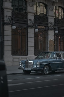 A vintage blue Mercedes-Benz is parked on a city street in front of a luxury boutique store with ornate ironwork and tall windows. The lighting is subdued, conveying a sense of elegance and timelessness.