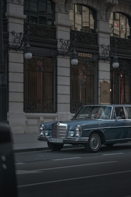 A vintage blue Mercedes-Benz is parked on a city street in front of a luxury boutique store with ornate ironwork and tall windows. The lighting is subdued, conveying a sense of elegance and timelessness.