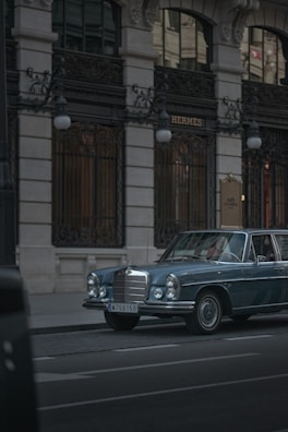 A vintage blue Mercedes-Benz is parked on a city street in front of a luxury boutique store with ornate ironwork and tall windows. The lighting is subdued, conveying a sense of elegance and timelessness.