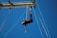 A person is suspended upside down in the air, secured by harnesses and ropes against a clear blue sky. Several ropes are attached to a metal structure above.