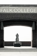 An outdoor area with a brick archway labeled 'Syracuse University Falk College'. Under the arch, there is a sculpture of a human figure placed on a pedestal. The background features a wooden bench and brick wall.