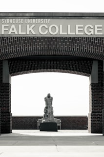 An outdoor area with a brick archway labeled 'Syracuse University Falk College'. Under the arch, there is a sculpture of a human figure placed on a pedestal. The background features a wooden bench and brick wall.
