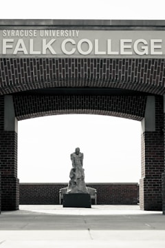 An outdoor area with a brick archway labeled 'Syracuse University Falk College'. Under the arch, there is a sculpture of a human figure placed on a pedestal. The background features a wooden bench and brick wall.