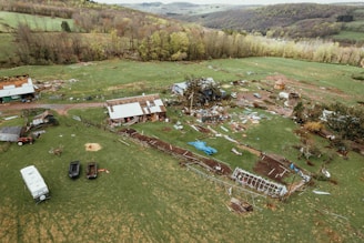 An aerial view of a rural landscape featuring a farm. The scene includes scattered debris from damaged buildings, including broken roofs and fallen trees, likely due to a storm. The area is surrounded by fields and woodlands, with a mix of green grass and brown patches of earth.