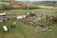 An aerial view of a rural landscape featuring a farm. The scene includes scattered debris from damaged buildings, including broken roofs and fallen trees, likely due to a storm. The area is surrounded by fields and woodlands, with a mix of green grass and brown patches of earth.