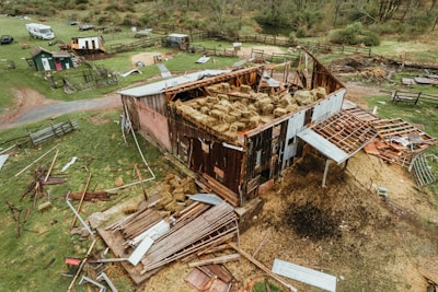 A partially collapsed barn surrounded by scattered debris and hay bales. The roof is significantly damaged, with sheets of metal and wooden planks strewn across the grass. There are multiple fenced enclosures and small structures in the background, along with some vehicles. The setting appears to be a rural farm area.
