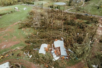 An aerial view of a landscape showing significant damage from a recent storm. The image depicts uprooted trees and debris scattered across a grassy area. A damaged building is visible, partially covered by broken branches and leaves. Several vehicles and trailers are present along a nearby road, with fields in the background.