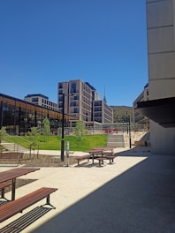 A modern architectural space featuring several multi-story buildings with extensive glass facades. There are neatly landscaped areas with grass and young trees. Wooden benches and tables are arranged in the open space, which is covered with light-colored pavement. A distant hill and a tall tower are visible in the background under a clear blue sky.