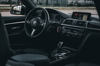 The interior of a black BMW car showcases a sleek and modern design with a leather steering wheel, digital dashboard, and control panel. The gear shift and infotainment display are prominently featured, exuding a sense of luxury and high technology.