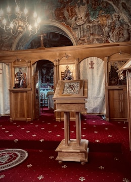 An ornate wooden lectern stands in the center of a richly decorated church interior, featuring religious icons. The space is adorned with intricate frescoes depicting various figures, illuminated by a chandelier. The floor is covered with a red carpet patterned with small white designs.