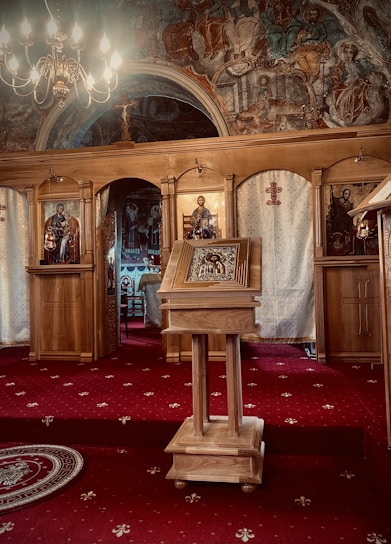 An ornate wooden lectern stands in the center of a richly decorated church interior, featuring religious icons. The space is adorned with intricate frescoes depicting various figures, illuminated by a chandelier. The floor is covered with a red carpet patterned with small white designs.
