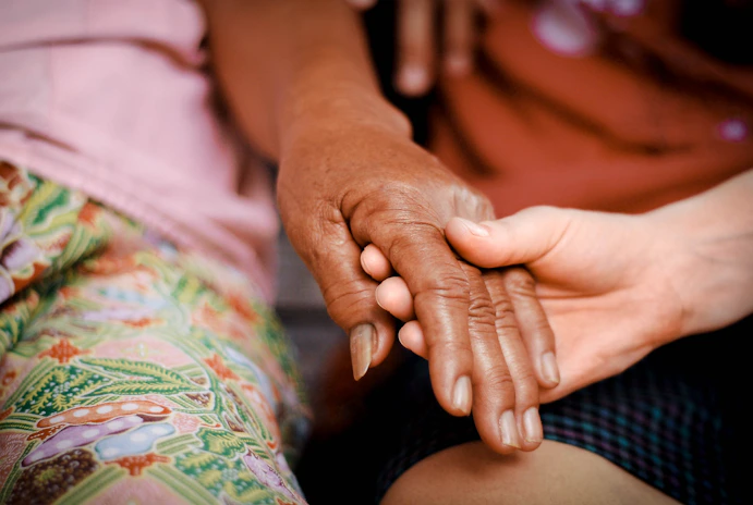 Two hands are touching gently, one older and darker-skinned, the other younger and lighter-skinned. The background includes vibrant, patterned fabrics.
