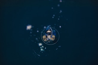 A person is floating underwater, wearing a mask and snorkel, with air bubbles surrounding them in a deep, dark aquatic environment.
