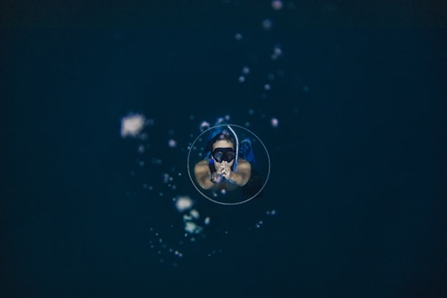 A person is floating underwater, wearing a mask and snorkel, with air bubbles surrounding them in a deep, dark aquatic environment.
