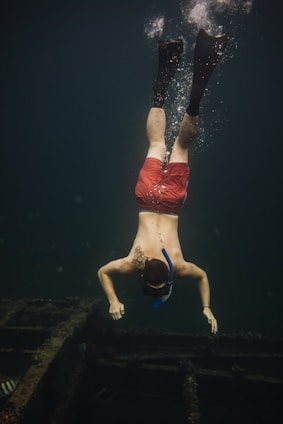 A person is diving underwater, wearing a snorkel and fins. They are swimming over a submerged structure, possibly a shipwreck, in the ocean. The water is dark, and the person's red shorts stand out against the deep greenish-blue surroundings.