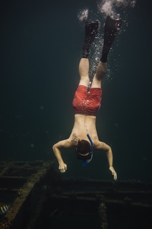 A person is diving underwater, wearing a snorkel and fins. They are swimming over a submerged structure, possibly a shipwreck, in the ocean. The water is dark, and the person's red shorts stand out against the deep greenish-blue surroundings.