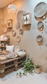 A cozy interior space features a variety of woven baskets hanging on a beige wall. The room includes a wooden pallet table with various decorative items such as textiles and ceramics. A small plant with green leaves is placed on the floor. Soft lighting and earthy tones create a warm and inviting atmosphere.