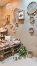A cozy interior space features a variety of woven baskets hanging on a beige wall. The room includes a wooden pallet table with various decorative items such as textiles and ceramics. A small plant with green leaves is placed on the floor. Soft lighting and earthy tones create a warm and inviting atmosphere.