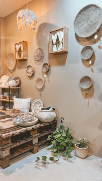 A cozy interior space features a variety of woven baskets hanging on a beige wall. The room includes a wooden pallet table with various decorative items such as textiles and ceramics. A small plant with green leaves is placed on the floor. Soft lighting and earthy tones create a warm and inviting atmosphere.