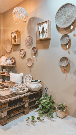A cozy interior space features a variety of woven baskets hanging on a beige wall. The room includes a wooden pallet table with various decorative items such as textiles and ceramics. A small plant with green leaves is placed on the floor. Soft lighting and earthy tones create a warm and inviting atmosphere.