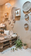 A cozy interior space features a variety of woven baskets hanging on a beige wall. The room includes a wooden pallet table with various decorative items such as textiles and ceramics. A small plant with green leaves is placed on the floor. Soft lighting and earthy tones create a warm and inviting atmosphere.
