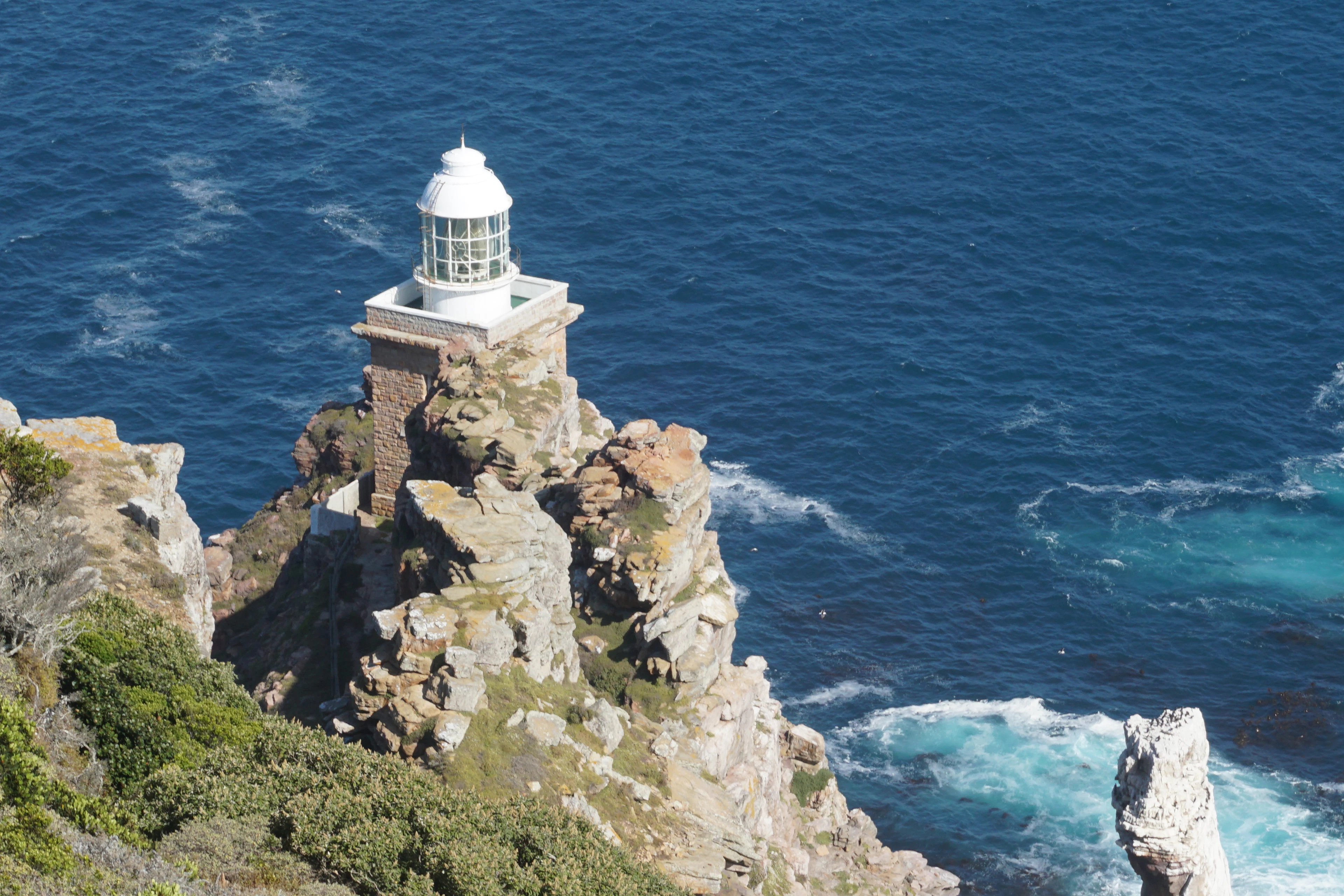 The white lighthouse at Cape Point standing on a rocky spur high above the ocean, deep blue water and turquoise surf breaking around the cliffs below it