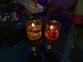 Two small decorative candles with flames are placed on a reflective surface. The candles are surrounded by small decorative items like confetti and balloons. The warm glow from the candles contrasts with the dark surroundings, indicating a festive or celebratory atmosphere.