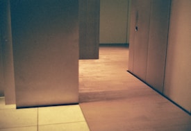 A minimalist interior space with geometric architectural elements. Light wood flooring is contrasted with soft beige walls, partially divided by vertical panels. The overall design is simplistic yet sophisticated, with a calm ambiance.