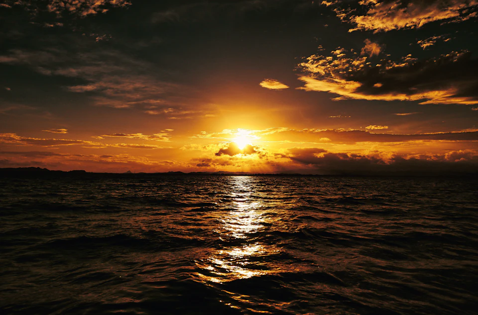 The image depicts a serene sunset over a body of water, with the sun setting behind the horizon and casting a warm glow across the sky. The deep blue ocean is dotted with small waves, reflecting the last rays of sunlight. The sky above is painted in shades of orange and yellow, creating a beautiful gradient effect as the sun sinks below the horizon.

In the foreground, there are several boats floating on the water's surface, adding depth to the scene. The image is taken from a high angle, providing an expansive view of the sunset over the ocean.