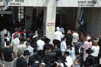 A crowd of people, primarily men and boys wearing traditional attire including fur hats and prayer shawls, are entering through a gateway. There is a separate entrance for women on the right. Several Israeli flags can be seen above the entrance.