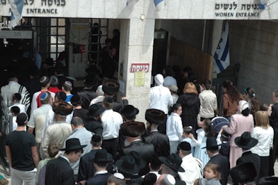 A crowd of people, primarily men and boys wearing traditional attire including fur hats and prayer shawls, are entering through a gateway. There is a separate entrance for women on the right. Several Israeli flags can be seen above the entrance.