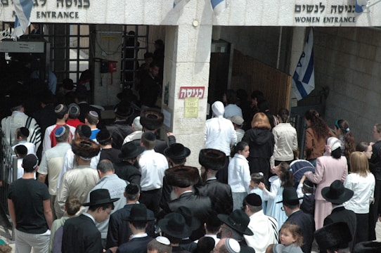 A crowd of people, primarily men and boys wearing traditional attire including fur hats and prayer shawls, are entering through a gateway. There is a separate entrance for women on the right. Several Israeli flags can be seen above the entrance.
