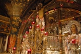 Ornate religious artwork dominated by a large crucifixion scene set against a richly decorated background, featuring a multitude of red votive candles. Icons and iconostasis panels adorned with silver and gold accents surround the crucifix. The ceiling and wall are covered with religious frescoes featuring angels and stars.
