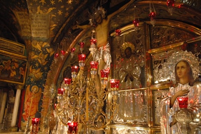 Ornate religious artwork dominated by a large crucifixion scene set against a richly decorated background, featuring a multitude of red votive candles. Icons and iconostasis panels adorned with silver and gold accents surround the crucifix. The ceiling and wall are covered with religious frescoes featuring angels and stars.