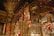 Ornate religious artwork dominated by a large crucifixion scene set against a richly decorated background, featuring a multitude of red votive candles. Icons and iconostasis panels adorned with silver and gold accents surround the crucifix. The ceiling and wall are covered with religious frescoes featuring angels and stars.